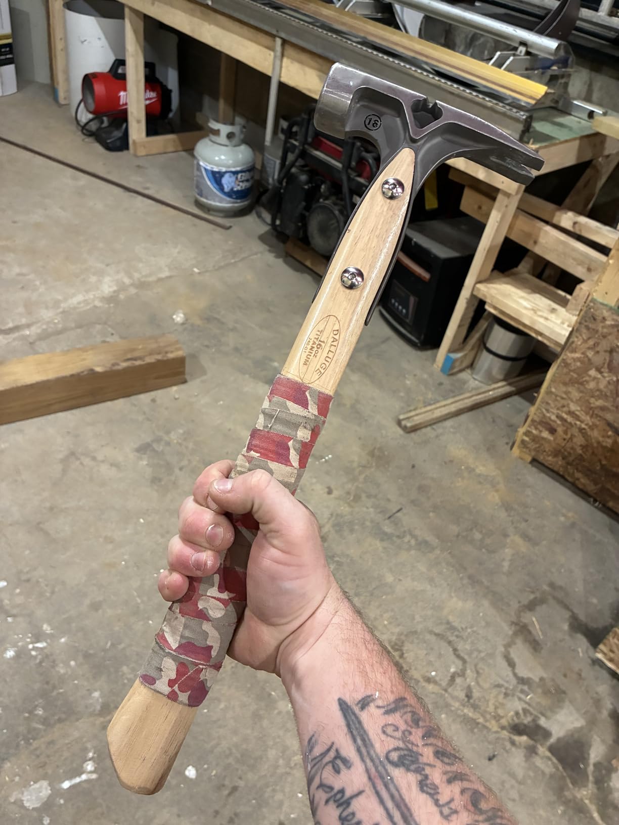 Owner holding a Dalluge 16 oz titanium hammer with camo tape wrapped around the hickory grip in a garage workshop