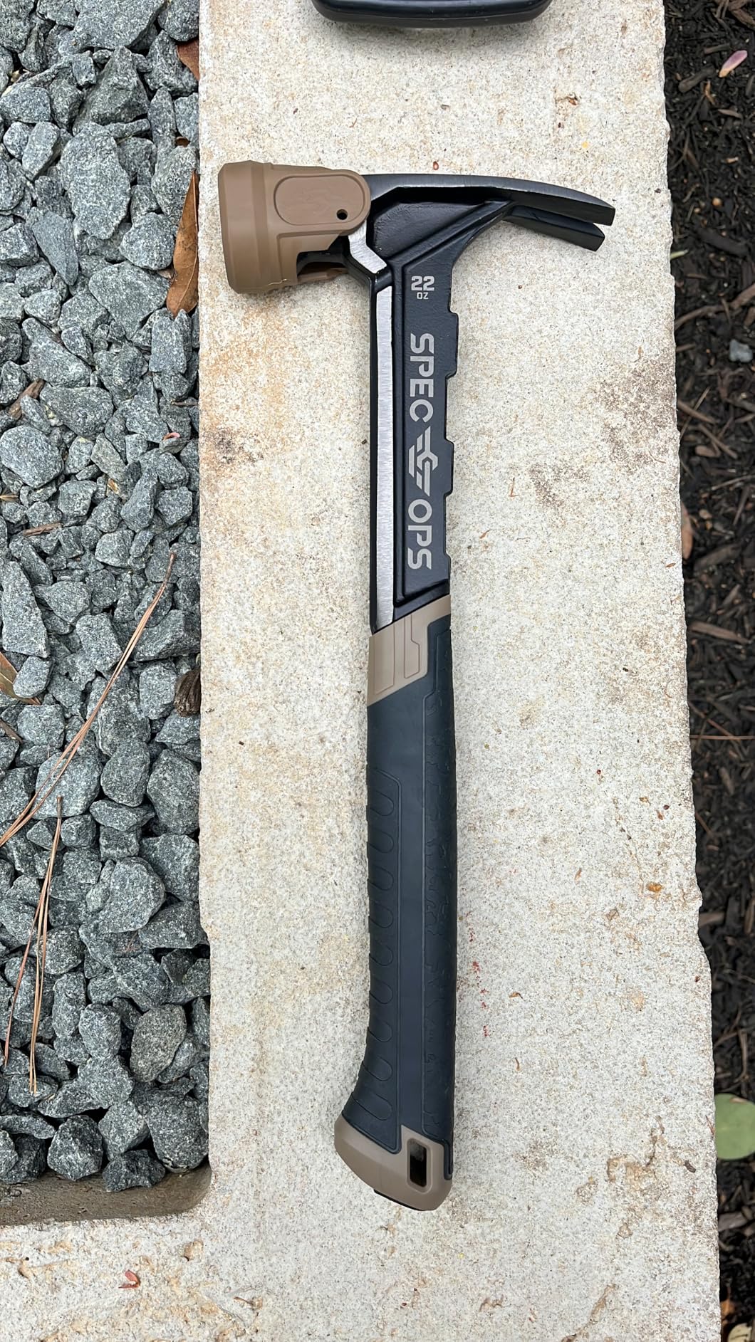 Black framing hammer with a tan face cap lying on a concrete ledge next to gravel